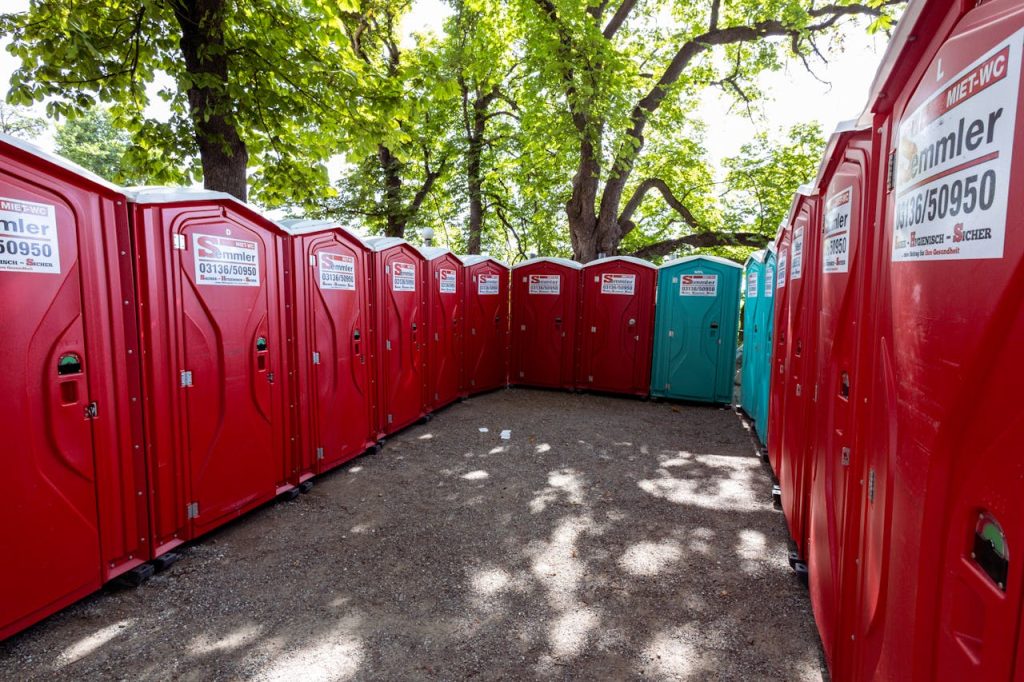 A line of red portable toilets under green trees in a sunny outdoor setting.