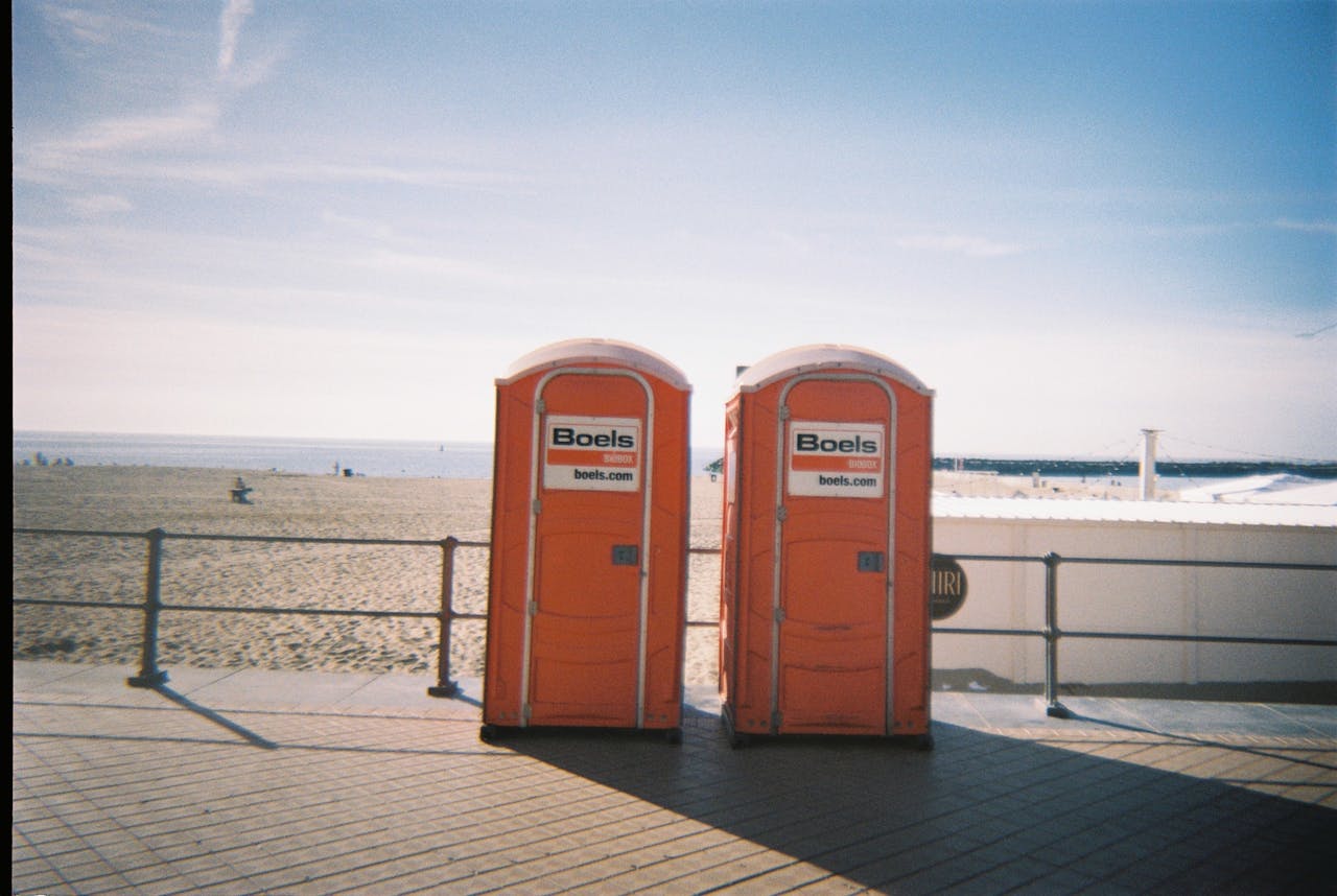Two orange portable toilets on a beach promenade with blue sky and sand.
